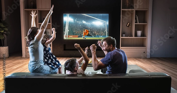 Obraz A family is watching a soccer moment on the TV and celebrating a goal, sitting on the couch in the living room.