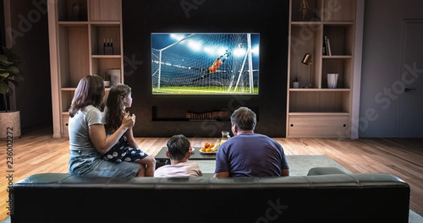 Obraz A family is watching a soccer moment on the TV and celebrating a goal, sitting on the couch in the living room.