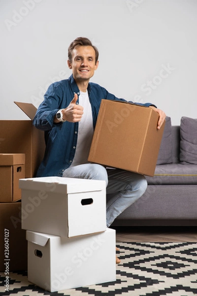 Fototapeta Image of happy brunet sitting on floor with cardboard boxes on background of gray sofa