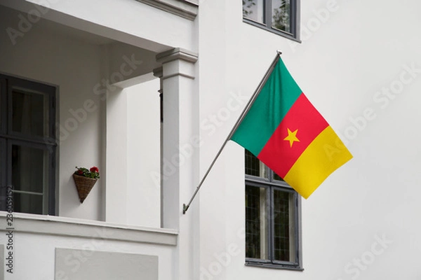 Fototapeta Cameroon flag hanging on a pole in front of the house. National flag waving on a home displaying on a pole on a front door of a building and raised at a full staff.