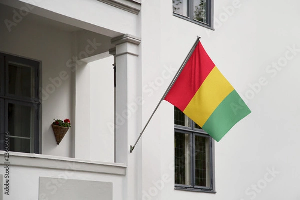 Fototapeta Guinea flag hanging on a pole in front of the house. National flag waving on a home displaying on a pole on a front door of a building and raised at a full staff.