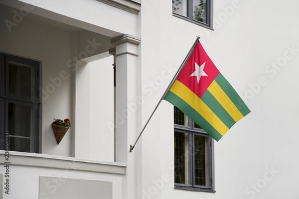 Fototapeta Togo flag hanging on a pole in front of the house. National flag waving on a home displaying on a pole on a front door of a building and raised at a full staff.