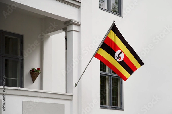Fototapeta Uganda flag hanging on a pole in front of the house. National flag waving on a home displaying on a pole on a front door of a building and raised at a full staff.