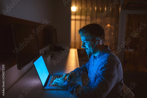 Fototapeta Young bearded Caucasian businessman typing on laptop and having earphones in ears while sitting in the office late at night.