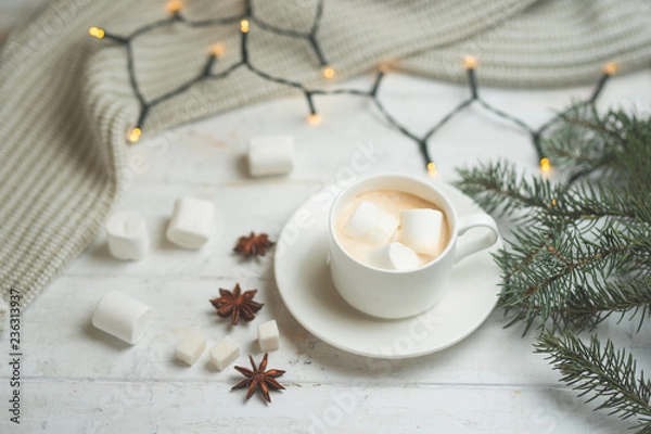 Fototapeta Coffee with milk, latte with cinnamon sticks and anise stars with white chocolate and marshmallow, with a branch of a Christmas tree on a bright white background, with lights from a garland.