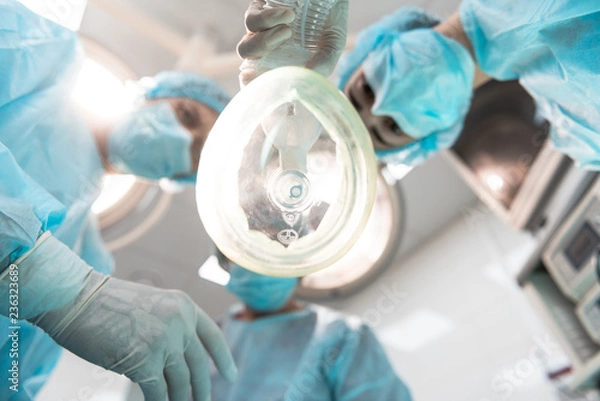 Obraz Widok z dołu trzech profesjonalnych lekarzy pochylających się nad pacjentem i noszących maski medyczne, trzymając jednocześnie inhalator znieczulający