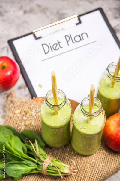 Fototapeta The diet plan is written on a white sheet on the tablet among the bottles of green smoothies, spinach and apples on a gray table with burlap. Weight loss concept. Top view