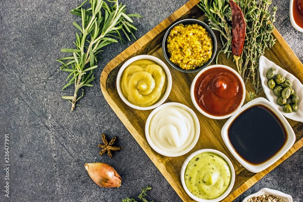 Fototapeta A set of sauces ketchup, mayonnaise, soy sauce, mustard, barbecue sauce, Dizhen mustard, horseradish on a dark stone background. Near spices pepper, rosemary, thyme, turmeric. Top view