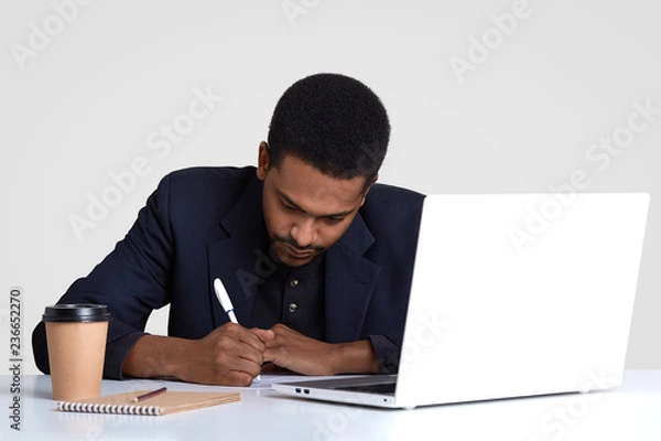 Obraz Busy male worker with dark healthy skin, writes down information in papers, holds pen, wears black suit, rewrites information from laptop computer, drinks coffee, isolated over white background