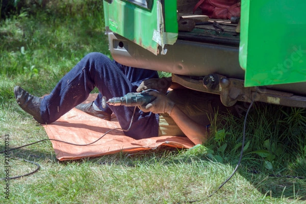 Obraz The mechanic lies under the car and repairs the body of a green minibus in the summer on the green grass outside.