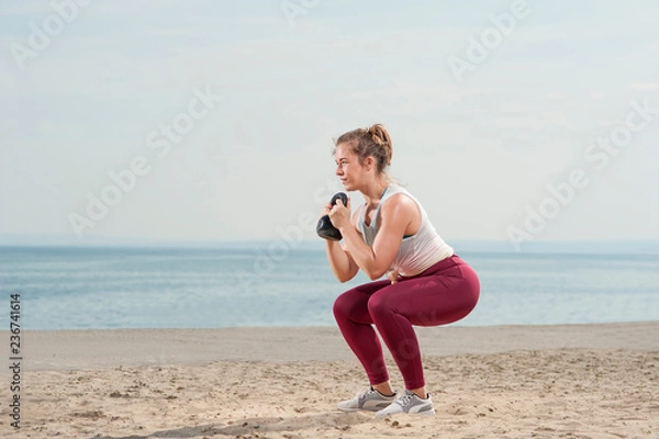 Fototapeta Young attractive woman dressed in pink leggings doing a squat with a kettle bell by the seaside. Fitness woman working out at the beach. Fitness Concept.