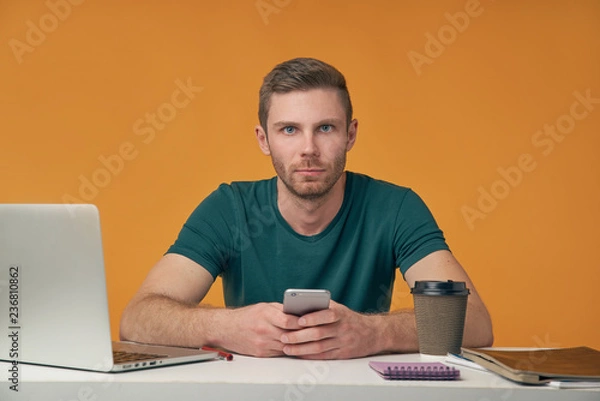 Fototapeta a young man with a beard in a t-shirt sitting at a Desk with a laptop and a Cup of coffee holding a smartphone and look carefully at the camera