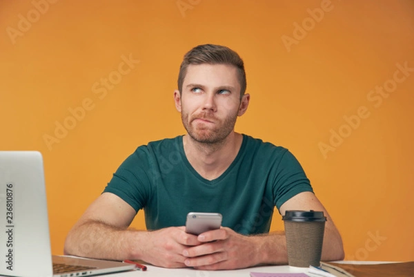 Fototapeta an attractive man in a t-shirt with a smartphone in his hands sitting at his Desk thinking and looking up. Isolated Studio portrait
