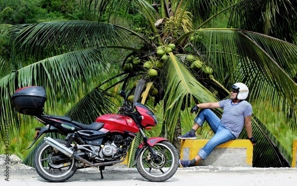 Fototapeta Biker with a motorcycle resting under a palm tree with coconats