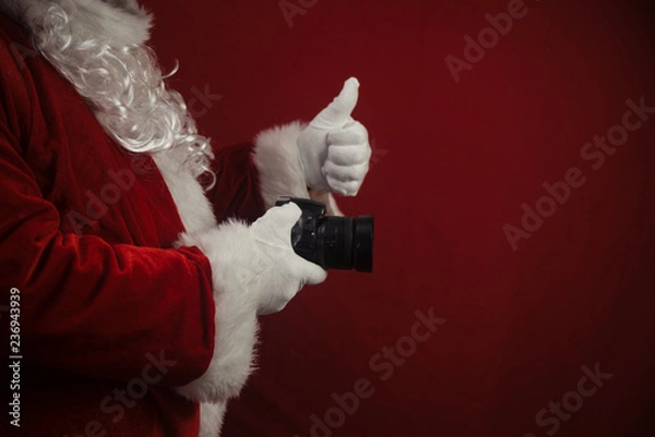 Fototapeta Santa Claus using DSLR camera taking images showing thumb up. Happy Christmas Evening and New Year celebration background. Fun loving creative costume time.