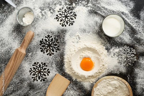 Fototapeta Christmas dark background with baking ingredients. Flour, egg, milk, rolling pin, sieve, snowflakes, spatula. Top view, place for text.