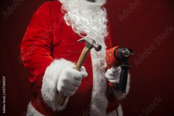 Fototapeta Santa Claus holding hammer and drill in hands busy preparing decoration. Closeup view of building creative ideas, taking job. Happy Christmas and New Year time renovation design background.