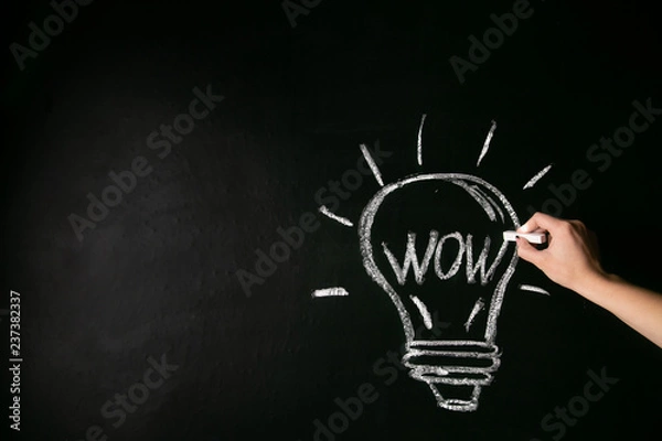 Fototapeta closeup female hand drawing a white chalk light bulb on a black board with the word wow