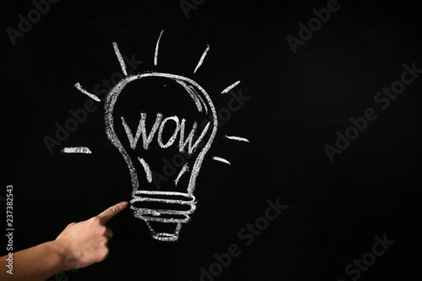 Fototapeta Closeup of female hand with raised index finger near chalked light bulb on black board with word wow