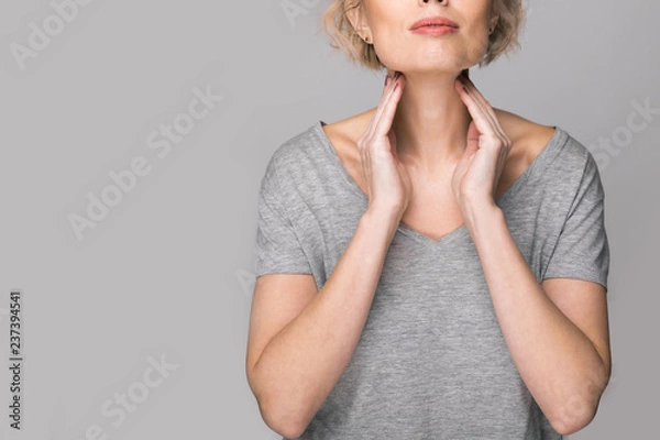 Obraz Female checking thyroid gland by herself. Close up of woman in white t- shirt touching neck with red spot. Thyroid disorder includes goiter, hyperthyroid, hypothyroid, tumor or cancer Health care.