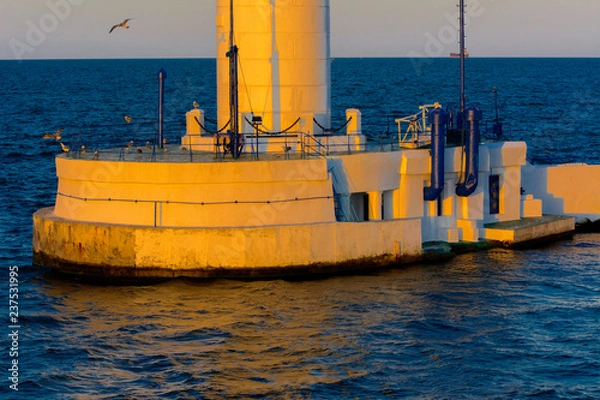 Fototapeta The lower part of the lighthouse at the entrance to  port on  background of a beautiful evening blue sky in soft colors. Summer seascape. White lighthouse with a red top. Black Sea. Seaport of Odessa