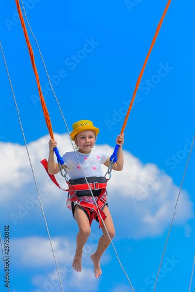 Obraz A little girl jumps high on a trampoline with rubber ropes against the blue sky and white clouds. Adventures and extreme sports. Concept of summer vacation, jumping.