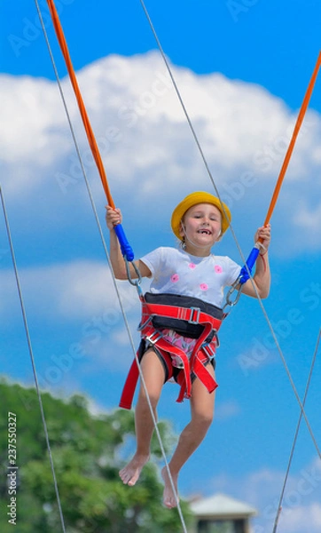 Obraz A little girl jumps high on a trampoline with rubber ropes against the blue sky and white clouds. Adventures and extreme sports. Concept of summer vacation, jumping.