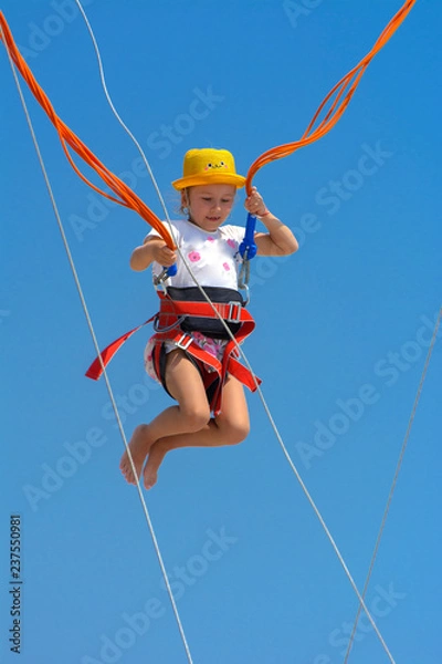 Obraz A little girl jumps high on a trampoline with rubber ropes against the blue sky and white clouds. Adventures and extreme sports. Concept of summer vacation, jumping.