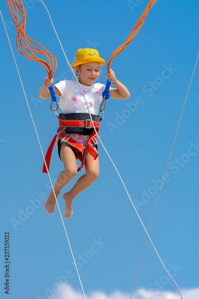 Obraz A little girl jumps high on a trampoline with rubber ropes against the blue sky and white clouds. Adventures and extreme sports. Concept of summer vacation, jumping.