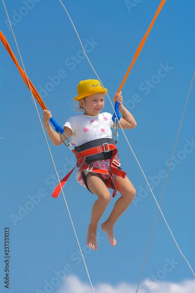 Obraz A little girl jumps high on a trampoline with rubber ropes against the blue sky and white clouds. Adventures and extreme sports. Concept of summer vacation, jumping.