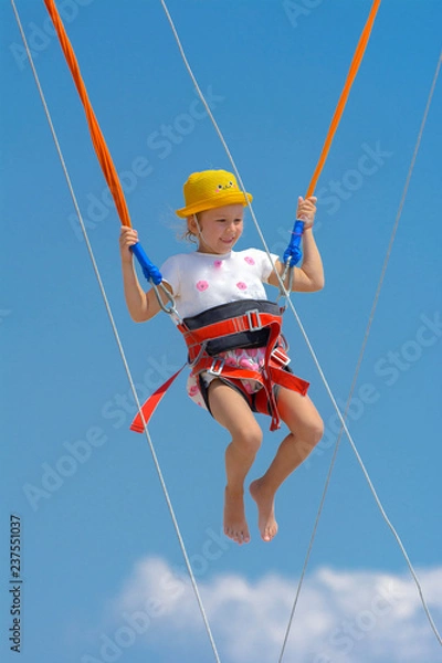 Obraz A little girl jumps high on a trampoline with rubber ropes against the blue sky and white clouds. Adventures and extreme sports. Concept of summer vacation, jumping.