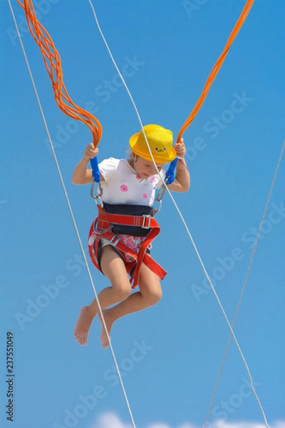 Obraz A little girl jumps high on a trampoline with rubber ropes against the blue sky and white clouds. Adventures and extreme sports. Concept of summer vacation, jumping.