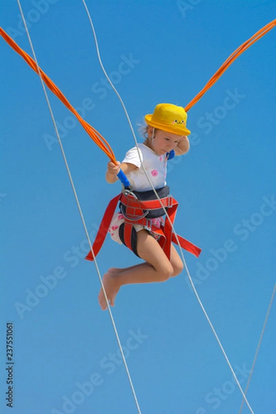 Obraz A little girl jumps high on a trampoline with rubber ropes against the blue sky and white clouds. Adventures and extreme sports. Concept of summer vacation, jumping.