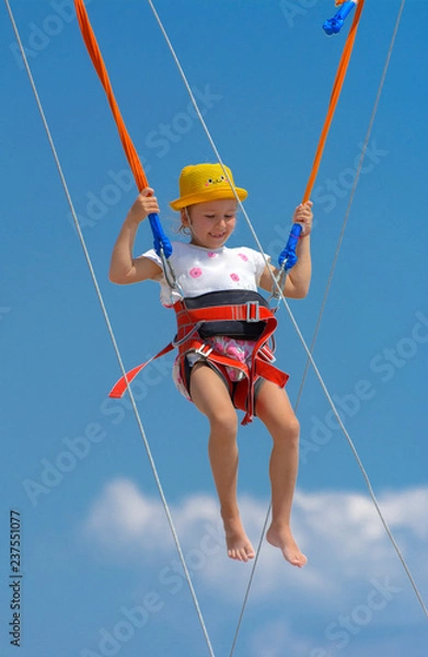 Fototapeta A little girl jumps high on a trampoline with rubber ropes against the blue sky and white clouds. Adventures and extreme sports. Concept of summer vacation, jumping.