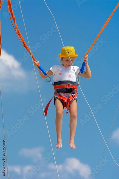 Obraz A little girl jumps high on a trampoline with rubber ropes against the blue sky and white clouds. Adventures and extreme sports. Concept of summer vacation, jumping.