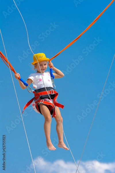 Obraz A little girl jumps high on a trampoline with rubber ropes against the blue sky and white clouds. Adventures and extreme sports. Concept of summer vacation, jumping.