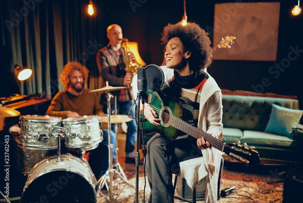 Fototapeta Mixed race woman singing and playing guitar while sitting on chair with legs crossed. In background drummer, saxophonist and bass guitarist.