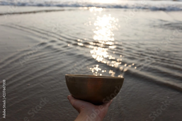 Obraz Tibetan singing bowl by the sea, in which the sun's rays shimmer. On the background are gentle waves and play of sunlight. Hand holds a bowl. Sound therapy. Healing. The music of the soul.
