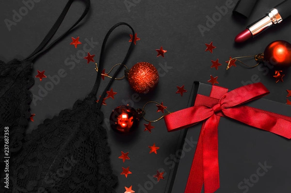 Obraz Czarne pudełko z czerwoną wstążką, koronkowa bielizna z biustonoszem, czerwona szminka, bombki, holograficzne konfetti na ciemnym tle, widok z góry flat lay. Niezbędne akcesoria damskie, prezent na nowy rok boże narodzenie