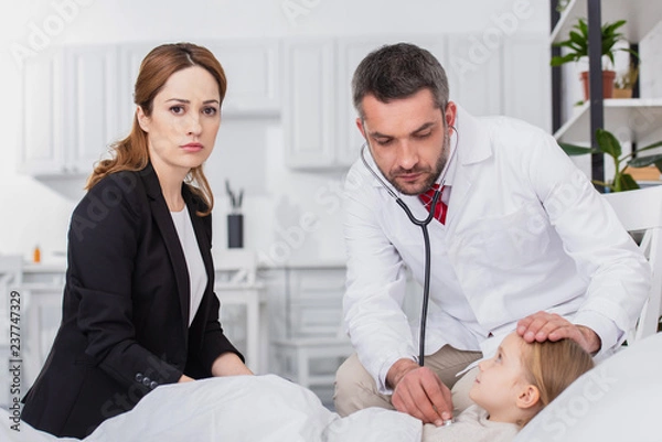 Obraz pediatrist in white coat examining sick kid with stethoscope at home, mother looking at camera