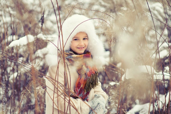 Fototapeta Little cute girl hide in high wet grass in snowy cold winter day playing with snowflakes