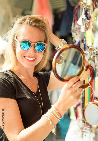 Obraz junge hübsche blonde frau auf dem markt in der sommer sonne in italien bei ihrer auslands reise. sie schaut sich sonnenbrillen, t-shirts, souveniers und taschen am markstand an und 