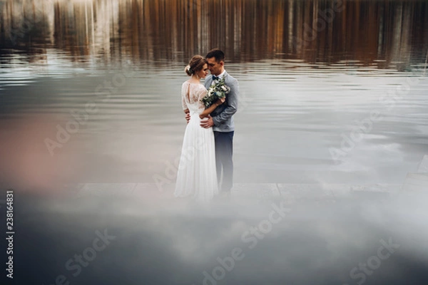 Fototapeta Panoramic view at youg couple bride in whire and groom in gray standing among lake, posing and embracing. Conceptual wedding at nature outdoor in autumn swasone. Wedding, family, love story concept.