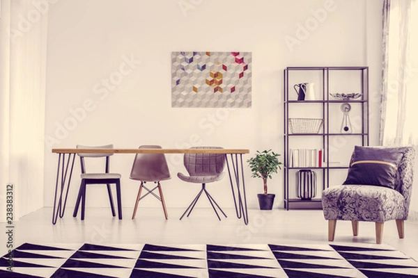 Fototapeta Pillow on grey armchair and geometric carpet in bright dining room with clock and poster on the wall and plant on the floor
