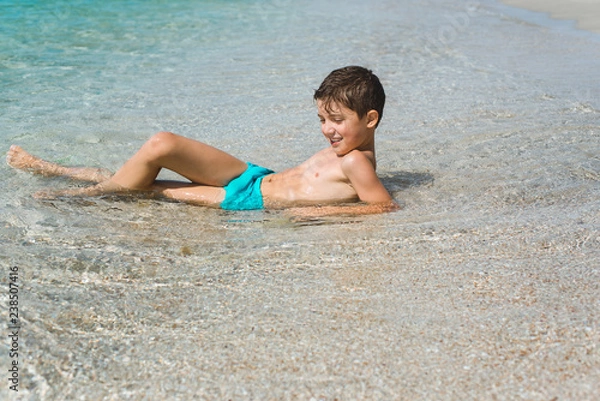 Fototapeta A cheerful kid on the beach lies in the clear sea water.