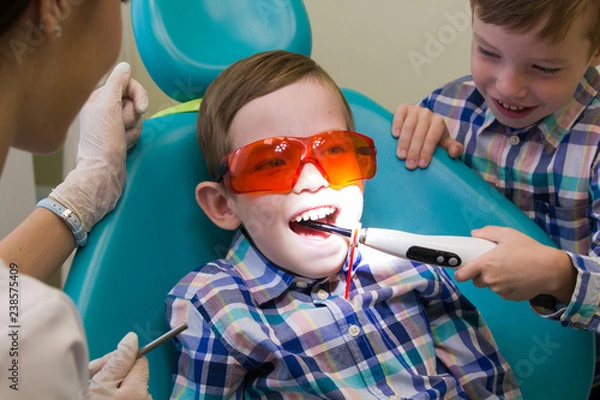 Fototapeta Reception at the dentistry. A little boy lays on the couch and his brother puts the lamp in his mouth