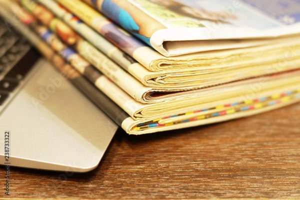 Fototapeta Newspapers and laptop. Pile of daily papers with news on the computer. Pages with headlines, articles folded and stacked on keypad of electronic device. Modern gadget and old journals, focus on paper 