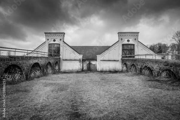 Fototapeta Vintage rural scene, dramatic front view of two black and white old rustic worn stone barn building farmhouse with dark sky. Two arched stone bridges in foreground.