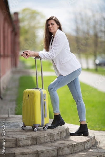 Fototapeta beautiful girl in a white jacket stands with a large yellow suitcase