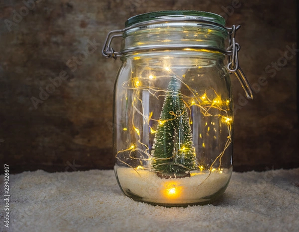 Fototapeta View of a jar with snow and a green Christmas tree decoration and Christmas lights on white snow and a wooden rustic background with copy space. Christmas interior decoration or design concept